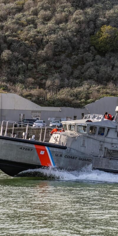 Operational U.S. Coast Guard boat cruising near seashore facility in daylight.