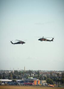 Two military helicopters hover above a city, offering a striking scene against the open sky.
